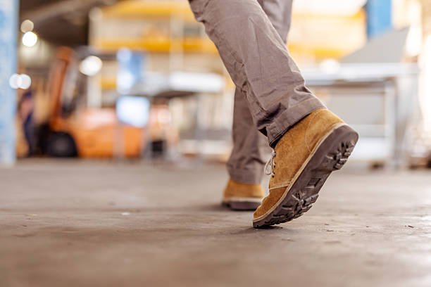 Worn leather boots stepping on a floor within a vibrant industrial setting, containing equipment and blurred background items. Captures movement and productivity in an active work location.
